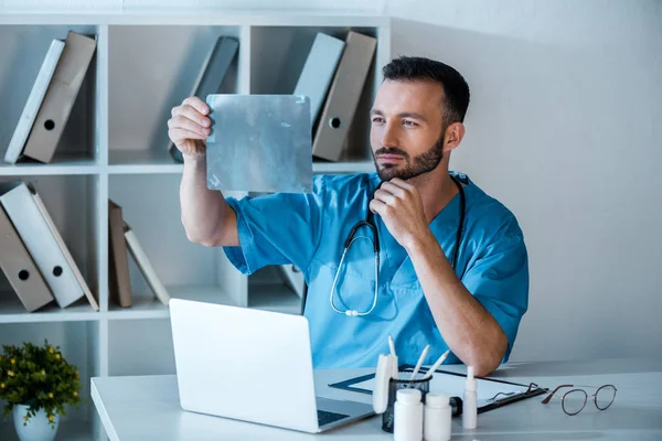 Ortopedista guapo mirando rayos X cerca del ordenador portátil en la clínica - foto de stock