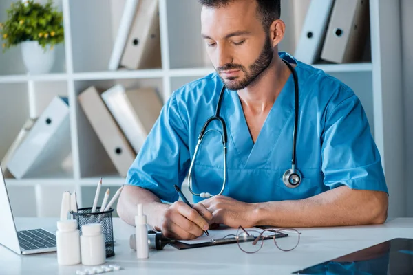 Handsome doctor writing prescription near laptop in clinic — Stock Photo