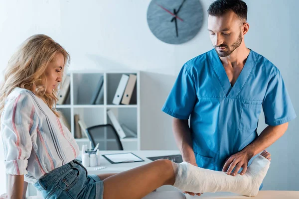 Красивый ортопед смотрит на сломанную ногу женщины — стоковое фото