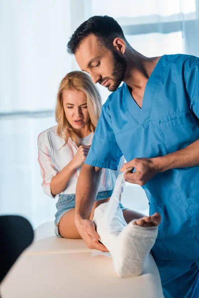 Bonito ortopedista colocando bandagem na perna fraturada da mulher assustada — Fotografia de Stock