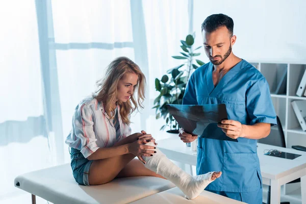 Handsome bearded orthopedist holding x-ray near injured woman in clinic — Stock Photo