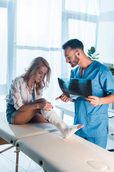 Handsome orthopedist holding x-ray and pointing with finger at injured leg of woman — Stock Photo