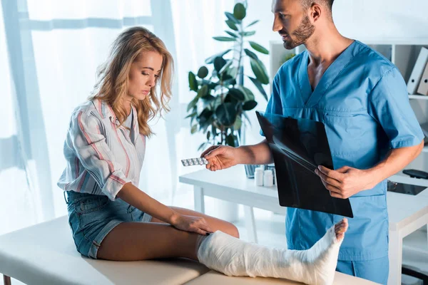 Handsome orthopedist holding x-ray and giving pills to injured woman — Stock Photo