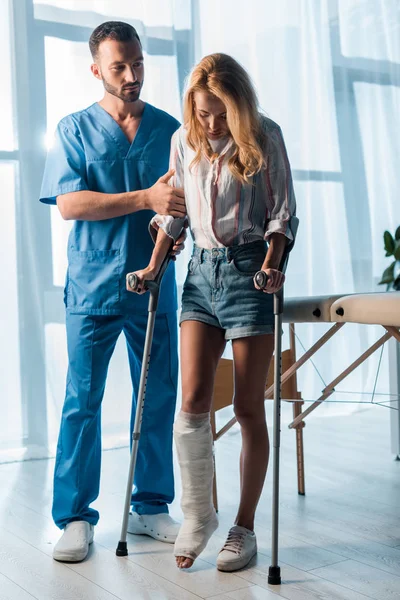 Красивый врач смотрит на раненую женщину, идущую с костылями — стоковое фото
