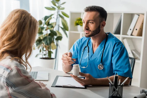 Selective focus of handsome doctor holding bottle with pills near woman in clinic — Stock Photo
