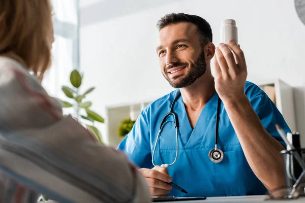 Enfoque selectivo del médico barbudo feliz sosteniendo la botella con pastillas cerca de la mujer - foto de stock