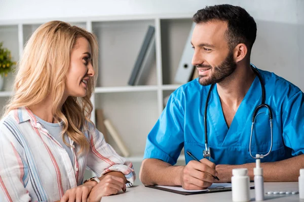 Enfoque selectivo de médico barbudo feliz sosteniendo pluma y mirando a la mujer atractiva en la clínica - foto de stock