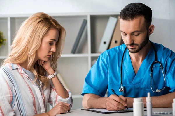 Handsome bearded doctor writing prescription to woman in clinic — Stock Photo