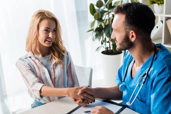 Selective focus of happy woman shaking hands with handsome doctor in clinic — Stock Photo