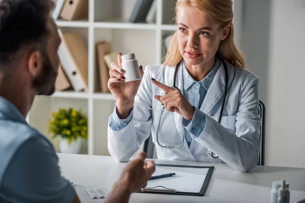 Selektiver Fokus des attraktiven Arztes, der mit dem Finger auf eine Flasche mit Pillen in der Nähe des Menschen zeigt — Stockfoto