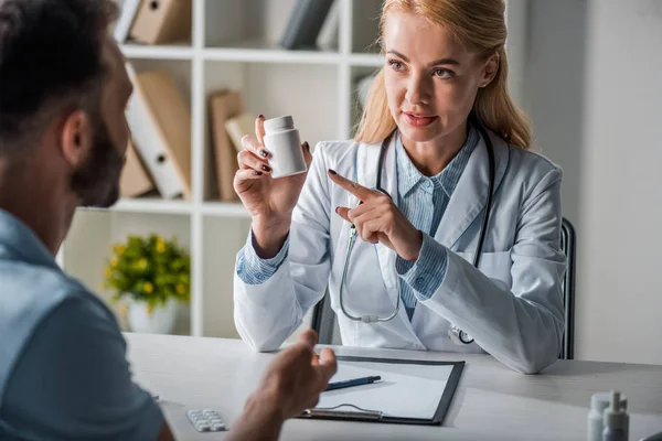 Fuoco selettivo del medico attraente che indica con il dito alla bottiglia con le pillole vicino all'uomo — Foto stock