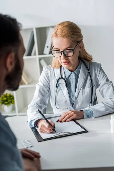 Selective focus of attractive doctor in glasses writing prescription near man — Stock Photo