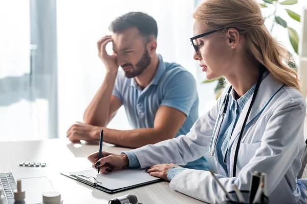 Selective focus of attractive doctor in glasses writing diagnosis near bearded man — Stock Photo