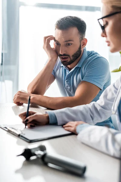Selective focus of shocked bearded man looking at doctor writing diagnosis — Stock Photo