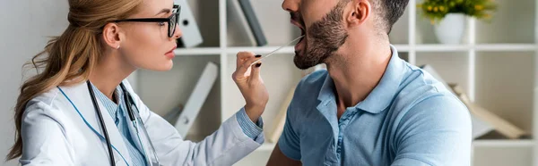 Plano panorámico de atractivo médico en gafas examinando hombre con espátula médica - foto de stock