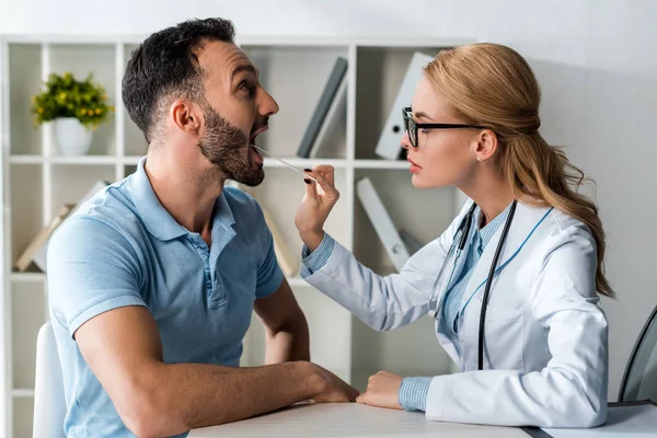 Side view of attractive doctor in glasses examining man with medical spatula — Stock Photo