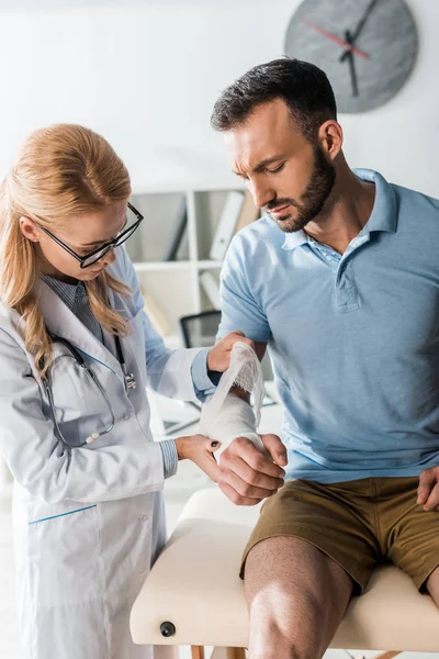 Atractivo ortopedista poner vendaje en la mano lesionada de hombre guapo - foto de stock