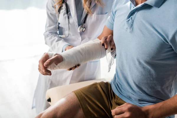 Vista recortada de ortopedista poner vendaje en la mano lesionada del hombre - foto de stock