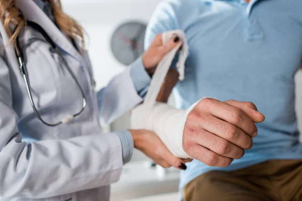 Vista recortada de ortopedista poner vendaje en brazo herido del hombre - foto de stock