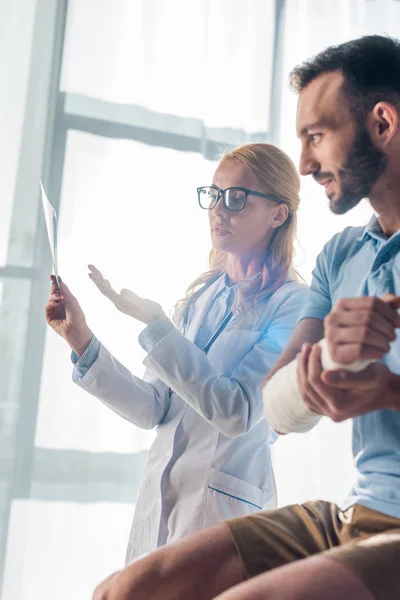 Selective focus of attractive orthopedist in glasses pointing with hand at x-ray near injured man — Stock Photo