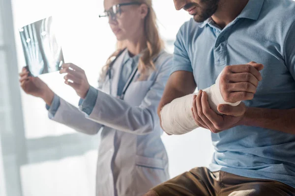 Selective focus of injured man near attractive doctor looking at x-ray — Stock Photo