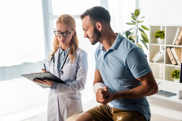 Attractive orthopedist holding clipboard near injured man — Stock Photo