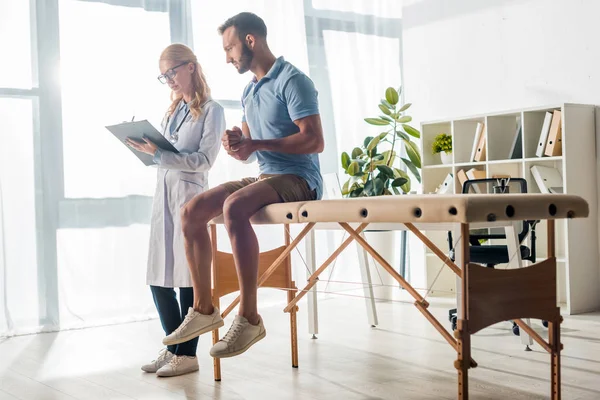 Beautiful orthopedist writing diagnosis near injured bearded man — Stock Photo