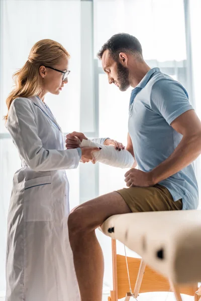 Selektiver Fokus attraktiver Orthopäden, die verletzte Hand eines bärtigen Mannes berühren — Stockfoto