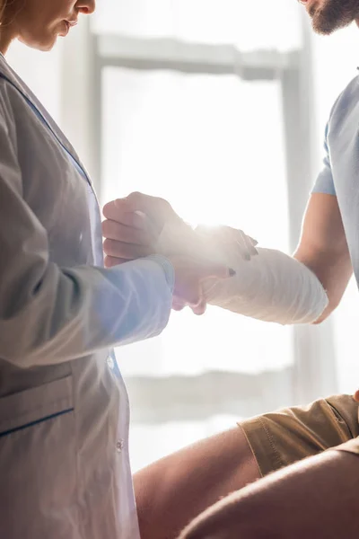 Vue recadrée de l'orthopédiste touchant la main blessée de l'homme à la clinique — Photo de stock