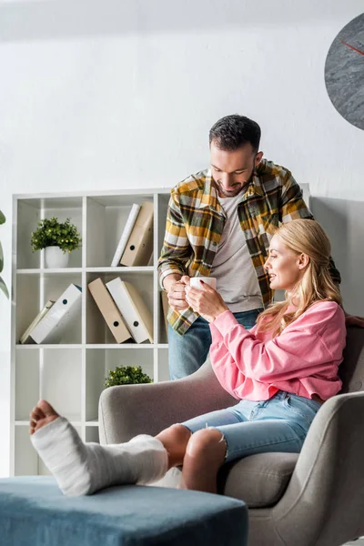 Handsome man giving cup of tea to injured woman at home — Stock Photo