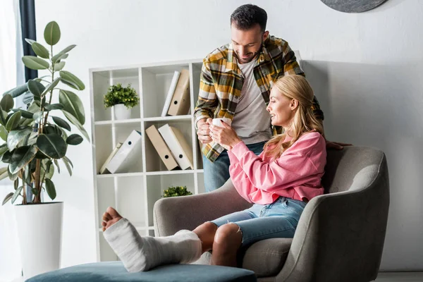 Homme barbu et beau donnant tasse de thé à la femme blessée à la maison — Photo de stock