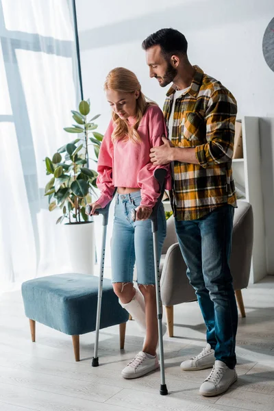 Hombre barbudo guapo de pie con mujer lesionada sosteniendo muletas en casa - foto de stock