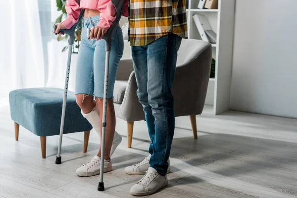 Vista recortada del hombre de pie con la mujer lesionada sosteniendo muletas en casa - foto de stock