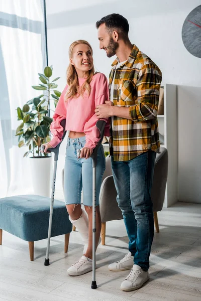 Hombre barbudo feliz mirando a la mujer lesionada sosteniendo muletas en casa - foto de stock