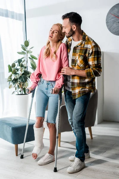 Hombre barbudo guapo mirando a la mujer lesionada sosteniendo muletas en casa - foto de stock