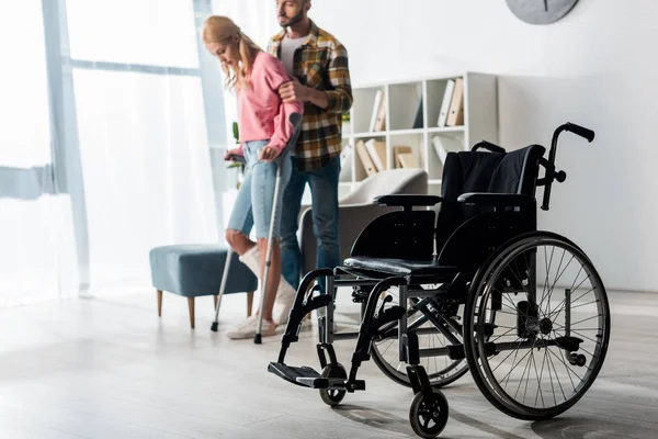 Selective focus of wheelchair near injured woman holding crutches while standing with man — Stock Photo