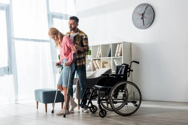 Wheelchair near injured woman holding crutches while standing with man — Stock Photo