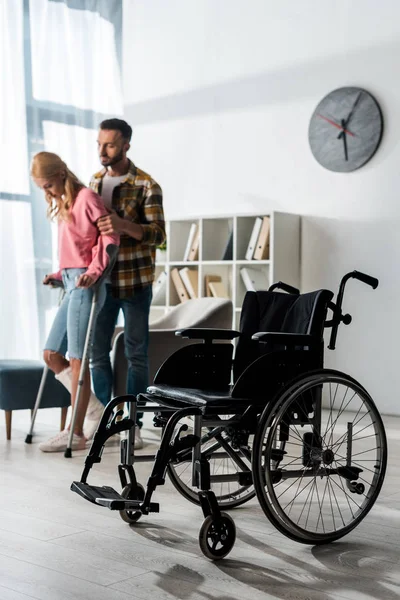 Selective focus of wheelchair near woman holding crutches while standing with man — Stock Photo