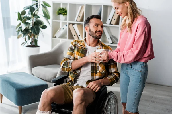 Femme gaie donnant tasse de thé à l'homme barbu en fauteuil roulant — Photo de stock