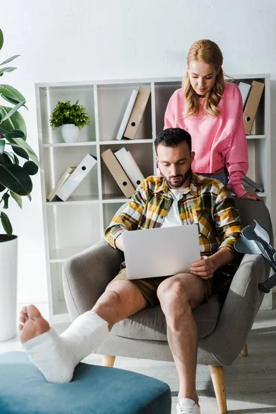 Attraktive Frau steht neben bärtigem verletzten Mann, der von zu Hause aus mit Laptop arbeitet — Stockfoto