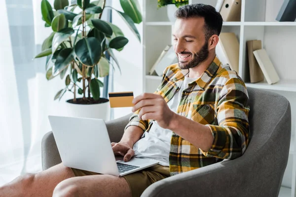 Uomo bello e sorridente in possesso di carta di credito mentre lo shopping online a casa — Foto stock