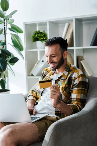 Sorrindo barbudo homem segurando cartão de crédito enquanto compras on-line em casa — Fotografia de Stock