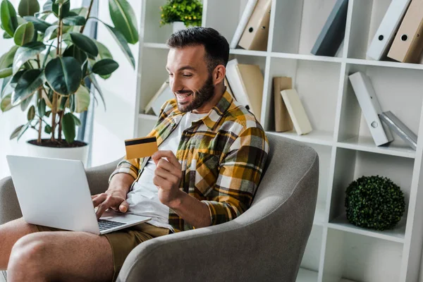 Hombre barbudo feliz celebración de la tarjeta de crédito mientras que las compras en línea en casa - foto de stock