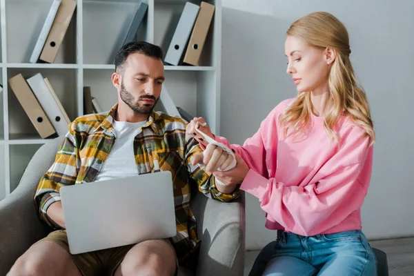 Donna premurosa mettere benda sulla mano ferita dell'uomo barbuto seduto con il computer portatile — Foto stock