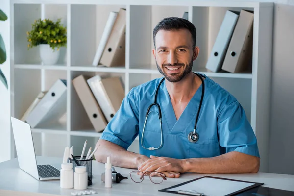 Felice medico barbuto seduto vicino a occhiali e laptop in clinica — Foto stock