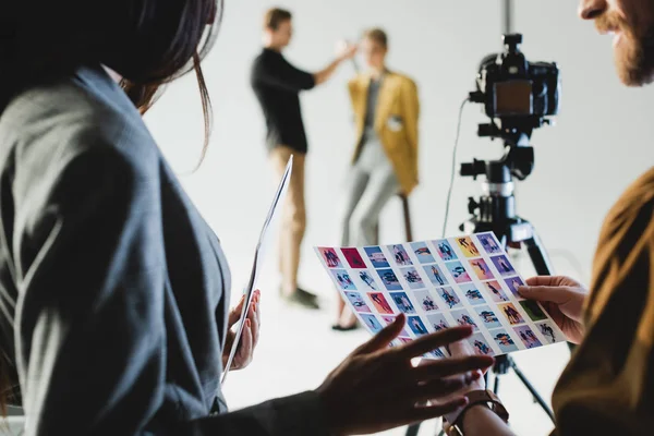 Selective focus of producer with photographer looking at references on backstage and hairstylist doing hairstyle to model on background — Stock Photo