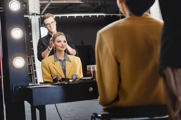 Messa a fuoco selettiva di parrucchiere facendo acconciatura a modello sorridente sul backstage — Foto stock