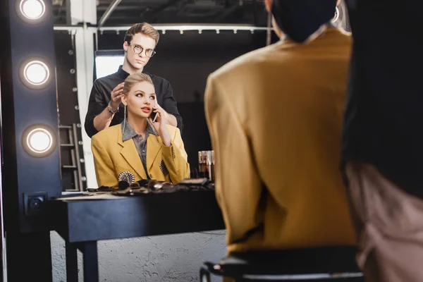 Foco seletivo do cabeleireiro fazendo penteado e modelo elegante falando no smartphone nos bastidores — Fotografia de Stock