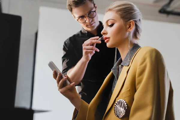 Vue à angle bas du maquilleur faisant du maquillage sur un modèle élégant avec smartphone dans les coulisses — Photo de stock