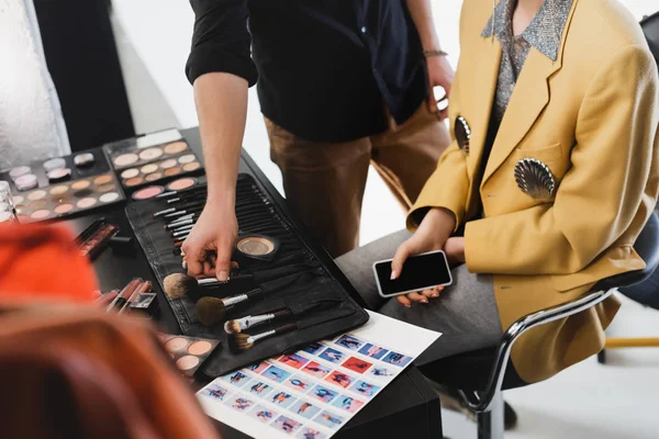 Vue recadrée de Makeup Artist prenant pinceau cosmétique et modèle tenant smartphone sur les coulisses — Photo de stock